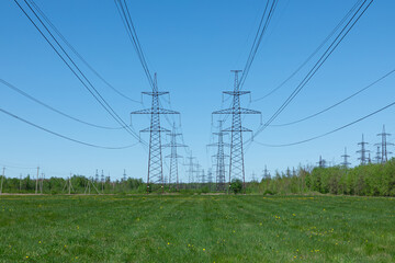 The towers of electric main in the countryside field on the background of blue sky and the forest with the wires in sunny summer or spring weather