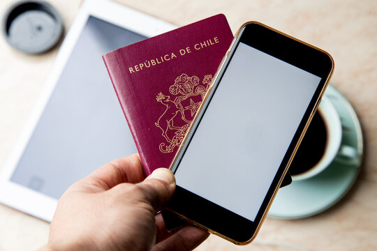 a top view of a hands holding a red Chilean passport and a black phone ,a table with a white tablet and a coffe 