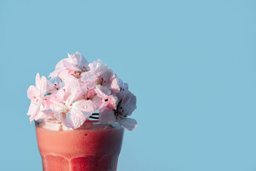The glass of smoothies is illuminated by sunlight. On top of the glass is a geranium flower in the form of a cap. The flower and drink are pink. The background is blue. There is space for text