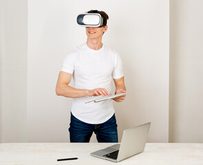 Male using virtual reality headset interacts with augmented things orienting in three dimensional space while sitting on his desk with keyboard and laptop on white background.