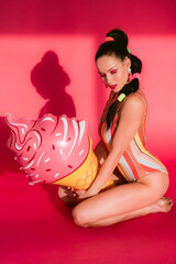 Obraz na płótnie Canvas Vertical full height body portrait slim busty body sexy seductive beautiful woman in retro swim suit look down isolated over shiny pink background sitting on knees with ice cream shaped baloon hand