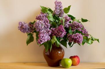 A beautiful bouquet of terry lilacs with green leaves in a brown ceramic vase and two apples.