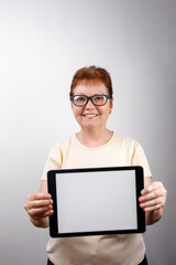 Senior woman in glasses shows a tablet on a white background in a light T-shirt. place for text, isolated