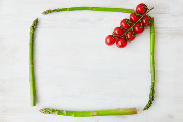 Square frame made with asparagus stems and decorated with a branch of cherry tomatoes in the corner. White rustic wooden background. Top view, flat lay. Copy space, space for text. Healthy food, diet