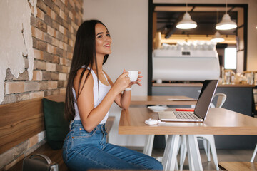 Attractive girl working on laptop in cafe. Beautiful girl using computer for work. Girl drinking cappuccino in cafe. Olnile work, freelancer. Female alone in cafe. Social distance