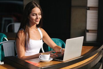Fototapeta premium Young business lady work on laptop and drinking a coffee. Beautiful girl sitting in cafe on terrace. Woman using phone and laptop outdoors