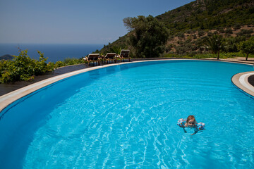 Little girl swimming in the swimming pool with a beautiful nature and sea view with trees and mountains.