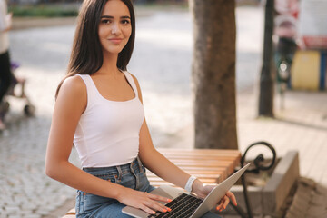 Attractive young woman use laptop outside on the bench in summer time. Woman working online outdoors. Alone. Beautiful brunette female. Student study