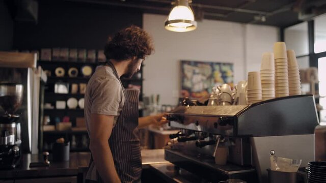 Young male chef in apron making fresh coffee in takeaway cup and giving to customer