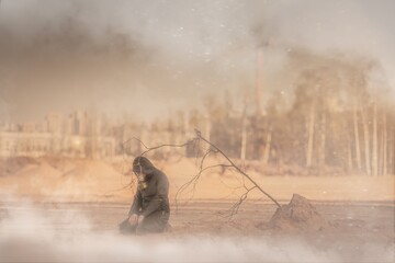 A man in a gas mask is kneeling in a desolate urban post-apocalyptic landscape. A dead tree sapling...