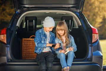 Slow motion of blond 12-aged girl and handsome 10-aged boy in jeans clothes which sitting in the trunk and looking on his smartphone