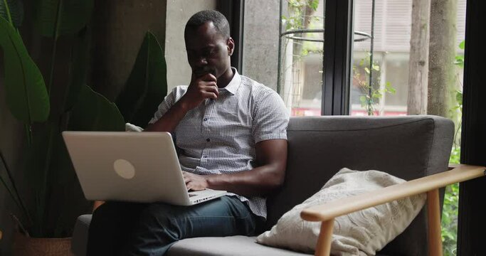 Close up of one young African businessman sitting in sofa typing on laptop in cafe office black young man using computer surfing internet thinking 