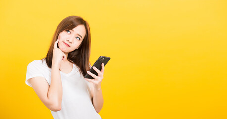 Fototapeta premium Asian happy portrait beautiful cute young woman teen standing wear t-shirt her using holding smart mobile phone and thinking looking to side isolated, studio shot yellow background with copy space