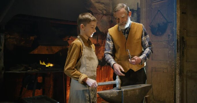 Caucasian senior grandfather blacksmith talking with cute teen grandson and demonstrating him producing of horseshoe. Grandpa teaching boy metal manufacturing in smithy.