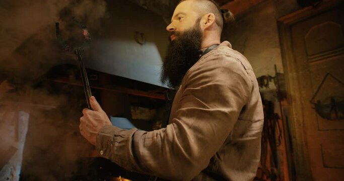 Young handsome male smith with long beard taking out molten glowing metal horseshoe from fire flame and looking at it with interest in antique smithy. Brutal forger working in workshop manufactory.