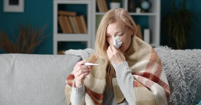 Sick blonde female covered with warm blanket, sneezing in tissue and measuring temperature at home. Mature lady suffering from flu while sitting on grey couch