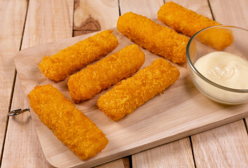 Pile of golden fried fish fingers with white garlic sauce placed on chopping board on wooden background