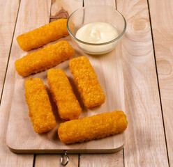 Pile of golden fried fish fingers with white garlic sauce placed on chopping board on wooden...