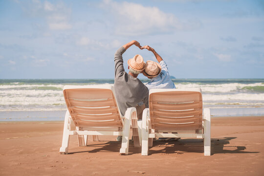 Rear view of senior couple sitting in deckchair on beach and Love making heart with hands. Love is everything,.Retirement age concept and love, copy space for text