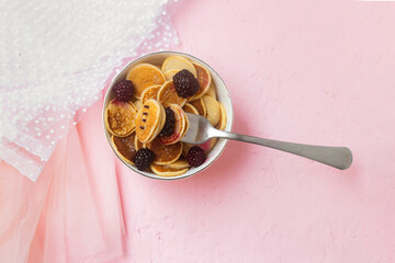 Mini pancakes with blackberry white dishes on a pink table. Lovely breakfast, morning light. A new trend dessert in social networks. Little Dutch poffertjes. Top view, flat lay.