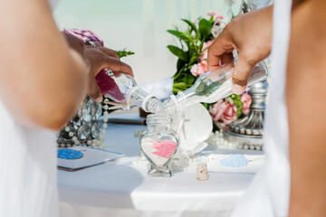 Bride and groom pouring colorful different colored sands into the crystal vase close up during symbolic nautical decor destination wedding marriage unity ceremony on sandy beach in front of the ocean 