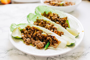 Delicious Asian Chicken Lettuce Wraps Served on a White Plate in a Kitchen Countertop with Romaine Lettuce and Lime Wedges.   