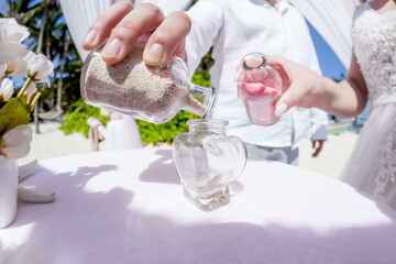 Fototapeta na wymiar Bride and groom pouring colorful different colored sands into the crystal vase close up during symbolic nautical decor destination wedding marriage unity ceremony on sandy beach in front of the ocean 