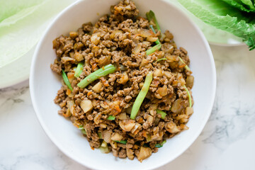 A White Bowl Filled with Ground Chicken and Long Scallions. Romaine Lettuce is in the Background. This ground meat dish is meant for asian lettuce wraps.