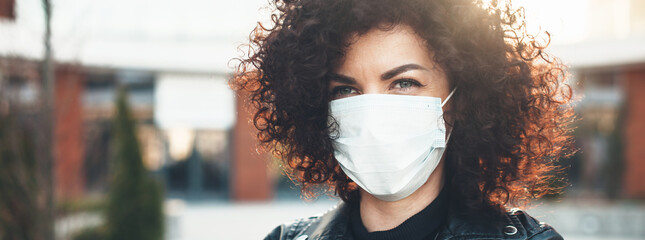 Close up photo of a curly haired woman wearing a protective mask outside and looking at the camera