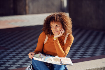 Young confused attractive mixed race girl with curly hair sitting on the stairs and holding map.
