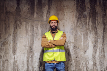 Young smiling attractive bearded worker in vest with helmet on head standing inside of building in construction process with arms crossed and looking at camera.