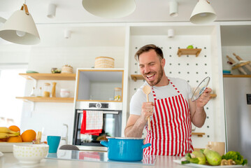 Young cheerful man wearing kitchen apron singing in the kitchen while preparing food