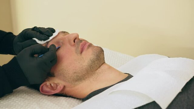 Close up of cosmetologist preparing man for the eyelash extension procedure. High quality close-up 4k slow motion video of a man laying on a couch during the esthetician lash treatment.