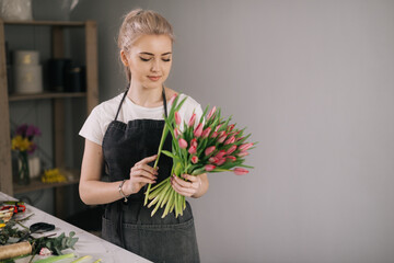 Cute young woman florist wearing apron making floral arrangement from fresh tulip at the table on...