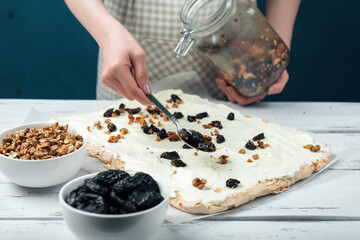 Woman spreads prune and nut filler on meringue cake on a white vintage wooden table.