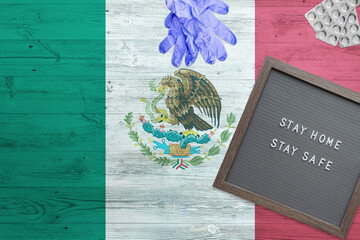 Mexico flag background on wooden table. Stay Home writing board, surgery gloves, pills with minimal national Covid 19 concept.