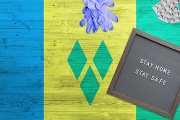 Saint Vincent And The Grenadines flag background on wooden table. Stay Home writing board, surgery gloves, pills with minimal national Covid 19 concept.