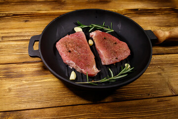 Raw pork steaks ready for preparation with rosemary, garlic and spices in cast iron grill frying pan on wooden table