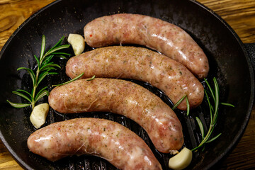 Raw sausages ready for preparation with rosemary, garlic and spices in cast iron grill frying pan on wooden table