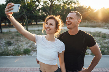 Young sporty couple in sportswear in urban park and taking selfie after workout on summer day on sunshine