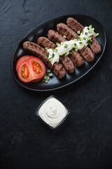 Fried cevapi or serbian skinless beef sausages topped with kaymak cheese, vertical shot on a black stone background with copyspace, top view