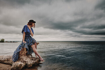 A girl in a blue dress and a black hat sits on the seashore on the street a cloudy sea of dark color.
