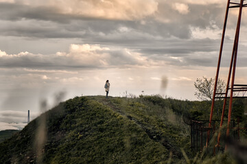 side view of a young girl on a hilltop with grass and cloudy sunset sky background near a cell tower. Hiking and outdoor concept.