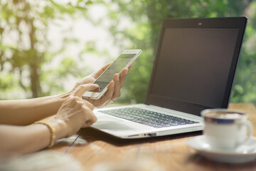 Obraz premium Woman using smartphone with laptop and a cup of coffee on the wooden table outdoor.
