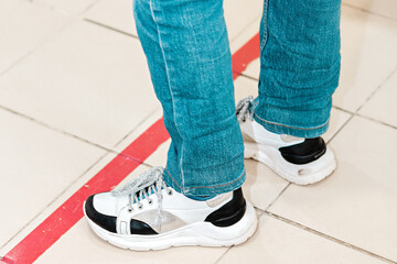 Person stand in red line, legsin shoes close-up. Attention line on the floor of the store to maintain social distance. Concept of the coronavirus pandemic and prevention measures