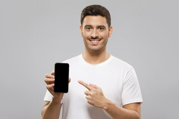 Young handsome man in white t-shirt holding smartphone, pointing to blank screen with finger, recommending mobile application, isolated on gray background with copy space