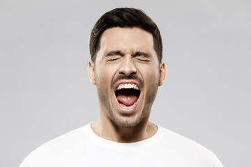 Headshot of man in white t-shirt screaming loudly with closed eyes, feeling strong anger and despair, suffering from trouble and exhaustion, isolated on gray background