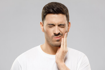 Young man in white t-shirt suffering from severe toothache, touching cheek with fingers, eyes closed because of strong pain, isolated on gray background