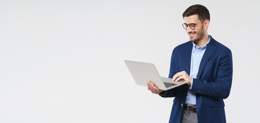 Horizontal banner of young handsome smiling business man holding laptop in hands, typing and browsing web pages, isolated on gray background with copy space - Powered by Adobe