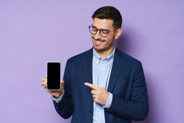 Young business man in jacket, shirt and eyeglasses holding smartphone with blank screen, copy space included, pointing to it, advising high quality finance app, isolated on purple background
